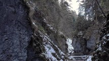 Backward Through Narrow Gap Between Rugged Cliffs With Wooden Bridge In Winter Forest. Pullback