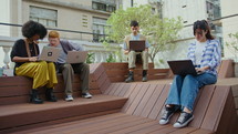 Group of gen Z business people in casual wear sitting on benches at public place in the city, using laptop and communicating, working remotely or studying online. Full shot
