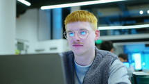 Young gen Z man wearing earrings, eyeglasses and casual outfit sitting at desk with coffee tumbler, using laptop and smiling, working in the office. Medium close-up handheld shot
