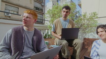 Business team of multi-ethnic gen Z coworkers sitting with laptops outside in the city, discussing ideas and plans, working together on summer day. PAN handheld shot
