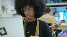 Young African-American businesswoman sitting at desk in open space office and working on laptop during the day. Medium close-up handheld shot
