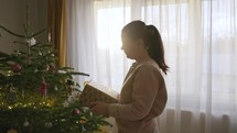 A Girl, Holding a Gift, Examines the Decorations Hanging on the Christmas Tree - Medium Close Up