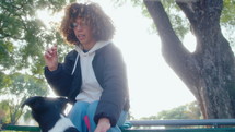 Young cheerful woman sitting on a bench in a public park, giving treats to a cute mixed-breed dog, smiling and petting him on a walk. Low angle shot
