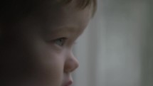 Closeup of the face of a young, cute boy, child inside watching the rain fall outside in cinematic slow motion.