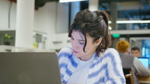 Young focused gen Z businesswoman in casual wear sitting at desk in the office, working with laptop, making notes and thinking. Medium close-up shot
