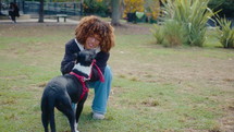 Young curly-haired woman crouching on a green lawn in the park, holding leash and petting her cute mixed-breed dog. Full shot, copy space

