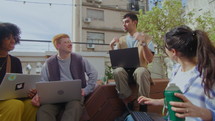 Company of multi-ethnic gen Z men and women sitting with laptops on wooden bench, having talk and smiling, working outdoors at public space in the city. Zoom shot
