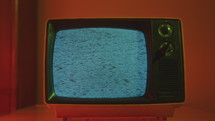 Old-fashioned analog TV with static noise on display and two dials on front panel in dark room with warm light. Zoom-off
