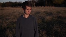 Young man, teenage boy standing in a field alone and watching the sunset or sunrise in cinematic slow motion.