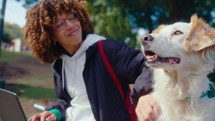 Young curly-haired woman smiling and petting adorable dog, then working on laptop, sitting in the park on a sunny day. Rack focus shot
