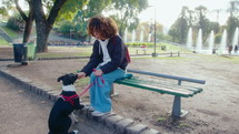 Young curly-haired woman sitting on bench in public park, treating lovely mixed-breed dog on a leash, having break during a walk
