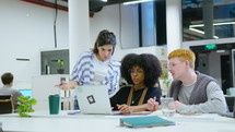 Young multi-ethnic male and female coworkers sitting at desk in open space office, working on laptop, having discussion about business project
