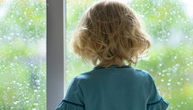 little girl with back to camera looks out a window on a rainy day