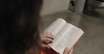 A convicted prisoner inmate wearing an orange prison uniform/jumpsuit sitting in his prison cell reading a bible.