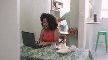 black female freelancer with curly short hair working online at laptop.Husband support and serve juice to his wife. Online education, work concept.