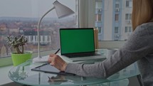 Woman writing notes on digital tablet on the desk laptop with green screen home office with big window view of cityscape