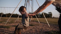 A father playing with his son at a park, pushing him on a swing in sunlight during sunset.