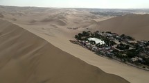 Aerial shot drone flies to left behind sand dune blocking desert city oasis Huacachina, Peru