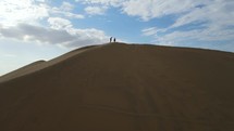 Aerial shot drone flies low ascending a giant sand dune from desert city oasis Huacachina, Peru meeting two hikers at the top for an extraordinary view of Ica