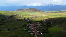 Aerial shot drone flies out from under cloud cover to a sun basked green field with farms and one hill towering over the landscape, dwarfed by large snow capped mountains in the distance