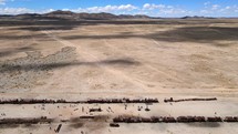Aerial shot drone flies left in wide shot of train cemetery in desert with mountains and white fluffy clouds in the distance contrasting the blue sky