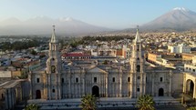 Aerial shot drone flies over Plaza de Armas through towers of Basilica Cathedral of Arequipa toward Misti Volcano and Chachani Volcano at dusk