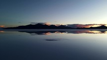 Aerial shot drone flies backwards low over salt flats covered in water with reflections of photographers and a mountain range in front of sunset