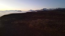 Aerial shot drone flies forward as camera pans down over red desert with snow capped mountains next to fog covered valley just before sunrise