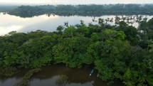 Aerial shot drone ascends from following canoe boat on black lagoon in middle of Amazon rainforest just before sunset to reveal an indigenous village and the rest of the lagoon