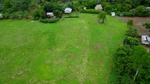 Aerial shot drone descending over open green field of indigenous village as hikers exit the jungle and enter the village in middle of Amazon rainforest