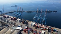 Aerial shot drone flies to the left and then orbits large cranes on loading dock with battleships on the next pier in coastal city on sunny day