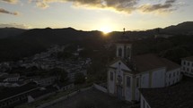 Drone flies in front of Igreja de Nossa Senhora das Mercês e da Misericórdia in Ouro Preto, Brazil toward sun setting behind hill