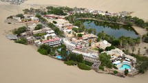 Aerial shot drone flies up and away from lake at oasis in middle of desert over couple sitting atop sand dune in Huacachina, Peru
