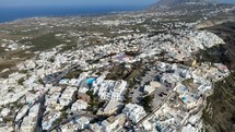 Aerial shot drone flies right and descends as camera pans left on iconic white buildings on the cliffs of Thera, Santorini