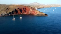 Aerial shot drone orbits to the right around Red Beach with two sailboats in bay in the south of Santorini