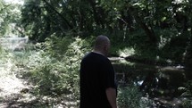 Middle aged Christian man in black shirt with beard in nature walking, hiking, meditating, praying contemplating in green, wooded area in trees with sunlight shining in cinematic, slow motion.