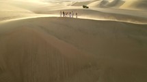 Aerial shot drone hovers as sandboarders wait at top of ridge as dune buggy drives behind them near desert oasis Huacachina, Peru