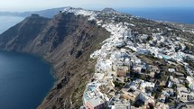 Aerial shot drone flies forward along cliffside of Thera, Santorini with iconic white buildings on sunny day