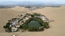 Aerial shot drone flies backwards from desert city oasis Huacachina, Peru over dune buggy parking lot