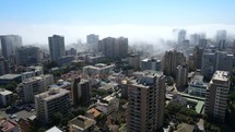 Aerial shot drone flies backwards from fog engulfing large buildings over dried up river with palm tree lined roads