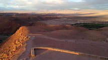 Aerial shot drone flies forward with back to sunset, over monument at viewpoint overlooking San Pedro and red desert mountains in Atacama