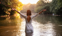 Woman in white dress with arms raised praising the lord, feeling free in a river during sunset.
