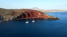 Aerial shot drone orbits to the left around Red Beach with two sailboats in bay in the south of Santorini