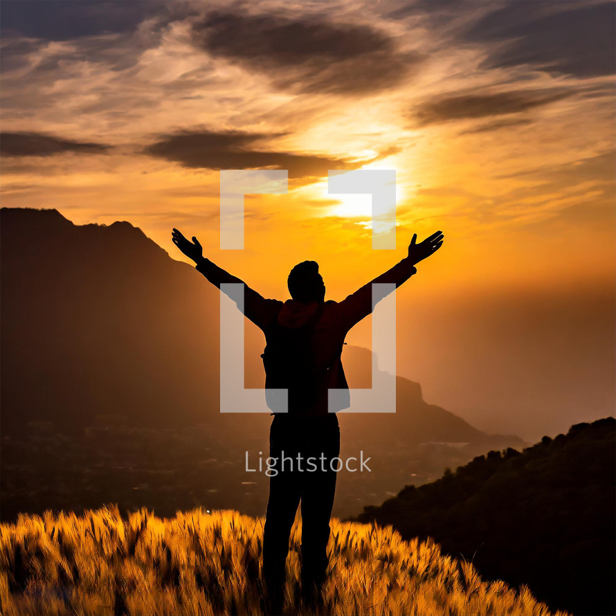 Silhouette of a man reaching his arms out on top of a mountain watching a sunrise with warm colors.