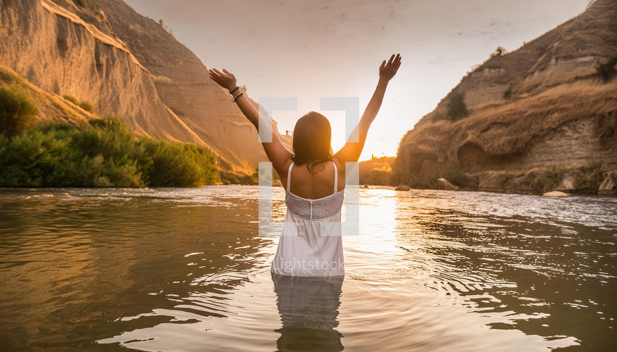 Woman in white dress with arms raised praising the lord, feeling free in a river during sunset.