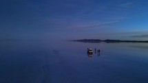 Aerial shot drone orbits to the left in wide shot around silhouettes of people and jeep on reflective water at sunrise