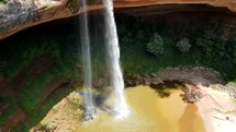 Aerial shot drone slowly ascends from pool at base of waterfall to the top with rainbow