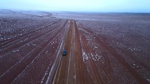 Aerial shot drone flies backwards following jeep through red desert with fresh snow on ground just before sunrise