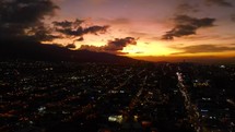 Aerial shot drone flies left while camera slowly pans right over lit up city at sunset