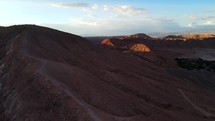 Aerial shot drone flies forward and to the left along path ascending hill in red desert at sunset